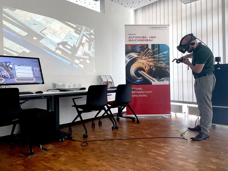 Mitarbeiter zeigt im Büro, wie virtuelles Schweißen mit VR-Brille funktioniert. Der digitale Vorgang ist auf der Wand links zu sehen. 