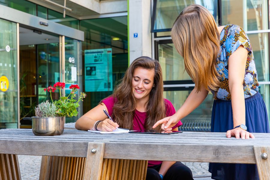 Zwei Studierende beraten sich. EIne sitzt und schreibt, die andere steht und deutet auf ein Tablet. 
