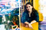 Eine Studierende in der Bibliothek, sie lehnt die Arme auf einen Stuhl und blickt in die Kamera, Im Hintergrundfährt ein Auto vorbei.