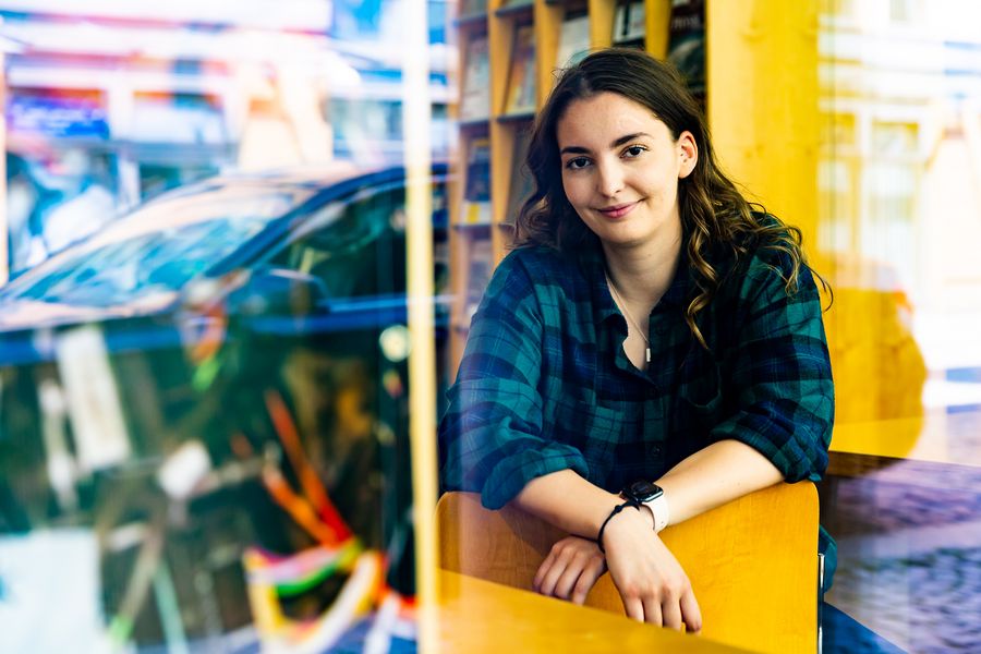 Eine Studierende in der Bibliothek, sie lehnt die Arme auf einen Stuhl und blickt in die Kamera, Im Hintergrundfährt ein Auto vorbei. 
