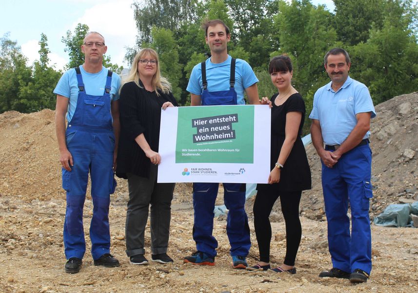 Foto: Gruppenbild mit 5 Personen. Diese halten ein Plakat mit der Aufschrift "Hier entsteht ein neues Wohnheim".