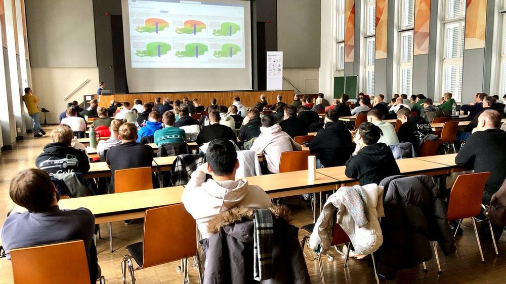 Gut gefüllte Aula. Die Studierenden sitzen an Tischen und verfolgen die Präsentation auf einer Leinwand. 