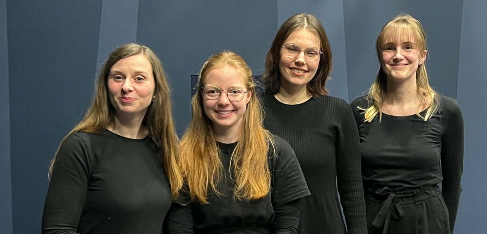 Vier Frauen in schwarzer Kleidung, die nebeneinander stehen und freundlich nach vorn schauen