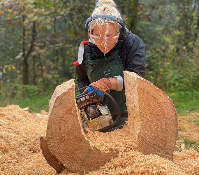 Studentin mit Kettensäge und Hörschutz bearbeitet einen Holzstamm