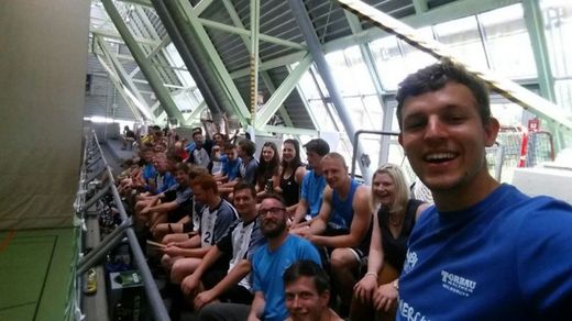 Foto: Hochschulsportteilnehmer sitzen auf einer Tribühne und lächeln in die Kamera.