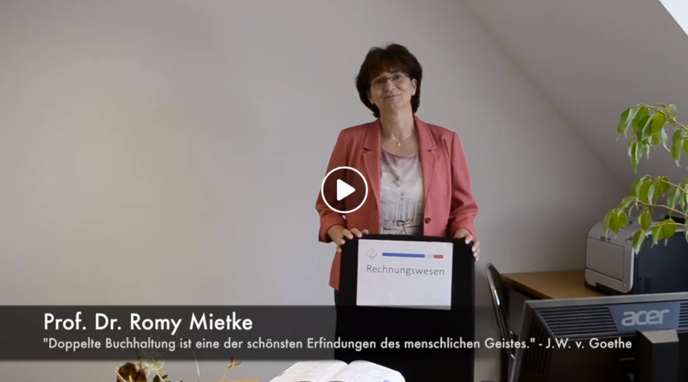 Videovorschaubild. Link zum Video. Frau Prof. Dr. Romy Mietke steht in ihrem Büro und zeigt ein Schild mit der Aufschrift Rechnungswesen.