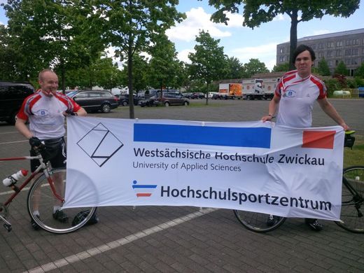 Foto: Zwei Herren mit Mountainbikes halten das Plakat mit Logo vom Hochschulsportzentrum hoch.