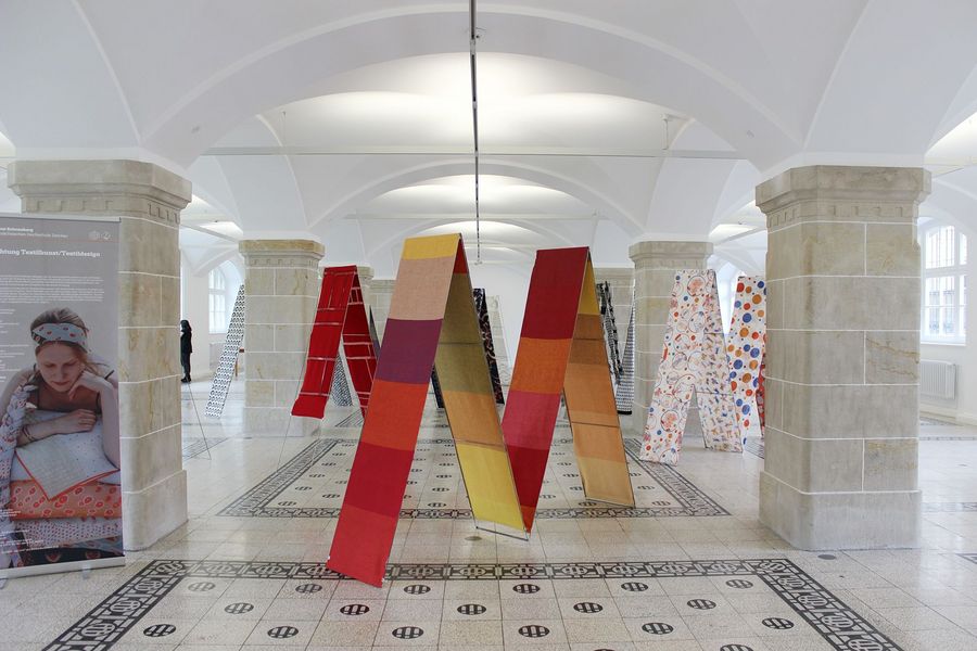 Foto: In einer Halle mit Säulen stehen aus textilem Material geformte große M (als Buchstabe).