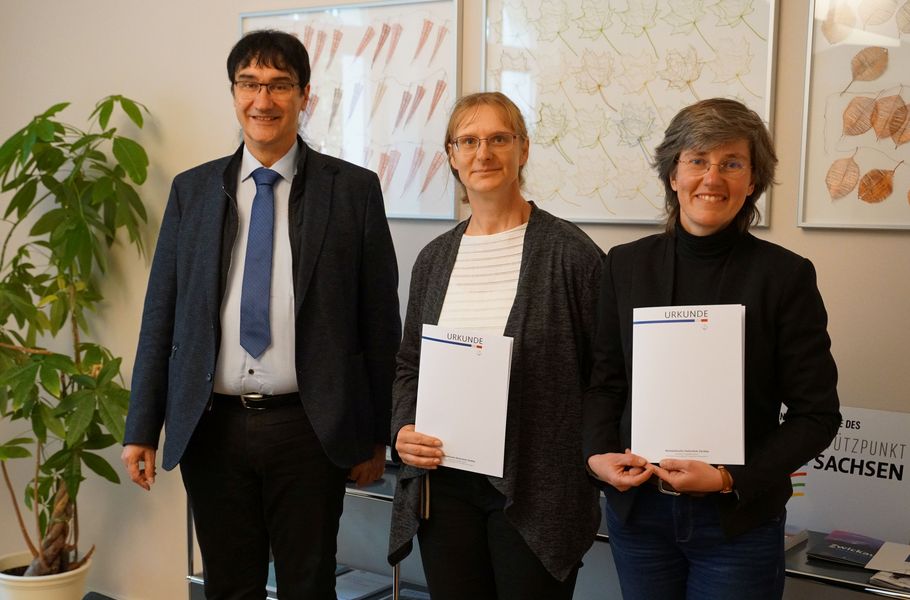 Foto: Neben einem Mann stehen zwei Frauen, die beide eine Urkunde in der Hand halten. 