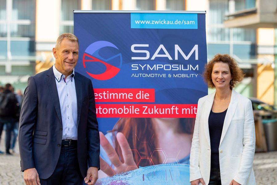 Ein mann steht links, eine Frau rechts vor einem Tranparent für SAM. 