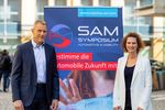Ein mann steht links, eine Frau rechts vor einem Tranparent für SAM.
