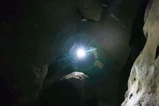 Foto: Outdoor in einer Höhle. Ein Teilnehmer mit Stirnlampe klettert durch eine Felsspalte.