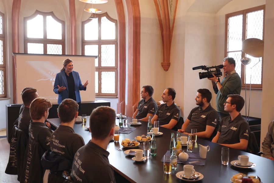Personen sitzen in einem festlichen Raum an einer gedeckten Tafel, Oberbürgerrmeisterin spricht, eine Kamera filmt das Geschehen. 