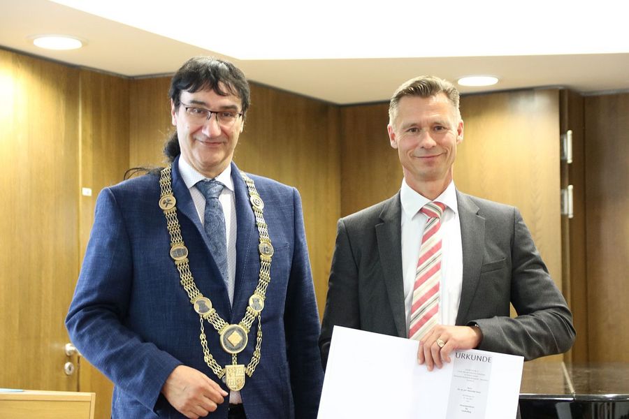 Ein Mann mit Amtskette steht links, ein Mann mit Urkunde in der Hand rechts daneben.