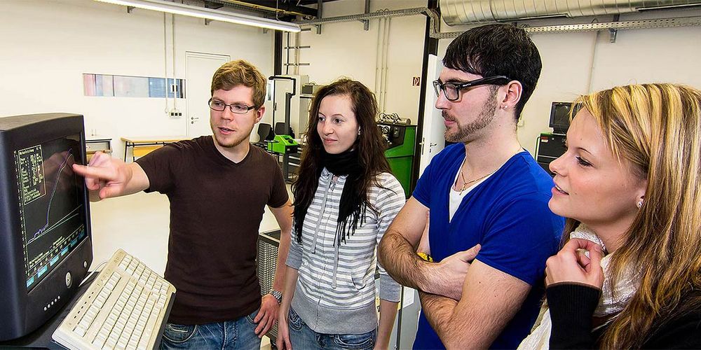 Foto: Ein Studierender zeigt drei weiteren Studenten eine grafische Darstellung mit zwei Kurven und Zahlenwerten auf einem Bildschirm.