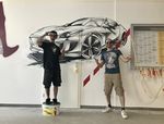 Foto: links steht ein junger Mann auf einem Farbeimer und hat ein Handy vor dem Gesicht, rechts steht ein junger Mann und präsentiert die mit einem Auto künstlerisch gestaltet ist. kauer Hauptbahnhofs