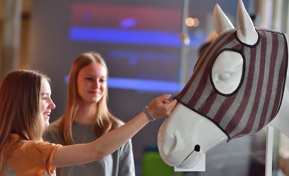 Foto: 2 Mädchen betrachten einen Zeckenkopfschutz auf einem Pferdekopfmodell. (Fotos: Wolfgang Schmidt, © vti - Verband der Nord-Ostdeutschen Textil- und Bekleidungsindustrie e.V.)