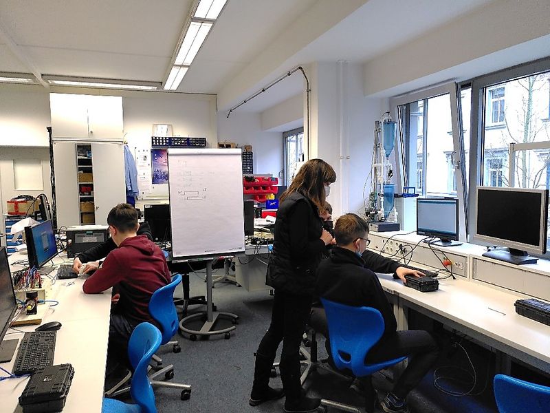 Foto: IN einem Labor sitzen 2 Schüler mit dem Rücken zur Kamera an Arbeitsplätzen, eine Frau steht zwischen ihnen.