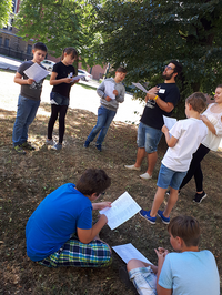 Foto: Eine Gruppe Jungs und Mädchen stehen und sitzen auf einer Wiese vor dem Agricola-Bau. Alle halten ein beschriftetes Blatt Papier in der Hand. Ein Mann ließt Ihnen etwas vor. 