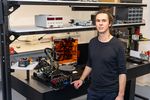 Foto: Junger Mann, schwarz gekleidet, steht vor Experimentiertisch, blickt in die Kamera.