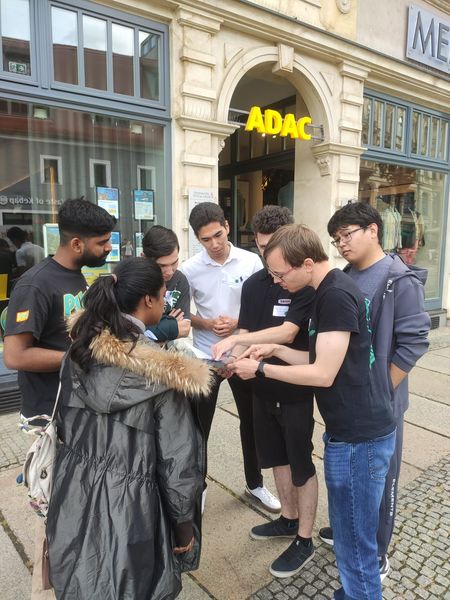 Eine Gruppe junger Menschen steht auf einem Gehweg vor einem historischen Gebäude mit einem gelben „ADAC“-Leuchtschild über dem Eingang. Sieben Personen sind im Kreis versammelt und schauen gemeinsam auf ein Tablet oder eine Karte, die in der Mitte gehalten wird. Eine Person trägt einen dicken Mantel mit Fellkragen, andere tragen T-Shirts oder leichte Kleidung. Die Stimmung wirkt konzentriert und kooperativ. Im Hintergrund sind Schaufenster mit Plakaten und Reflexionen zu sehen.