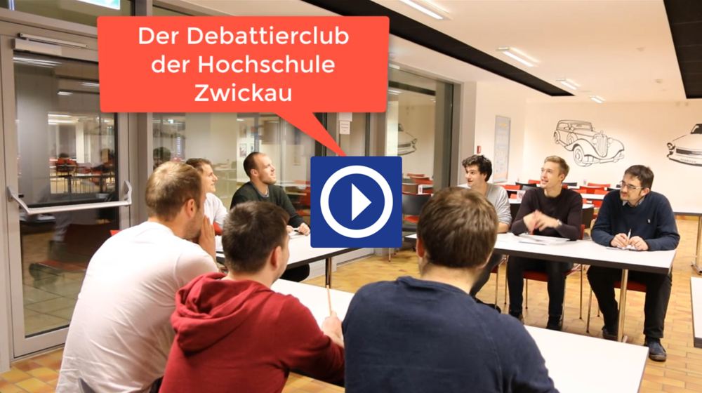 Videovorschaubild. Link zum Video Debattierclub. Eine Gruppe Studierender sitzt an einer o-förmigen Tischanordnung zusammen und debattieren miteinander.