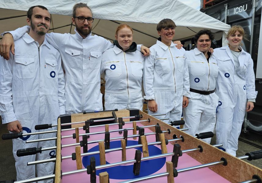 Kickertisch im Vordergrund dahinter sechs Studierende in weißen Arbeitsanzügen, welche Arm in Arm nebeneinanderstehen