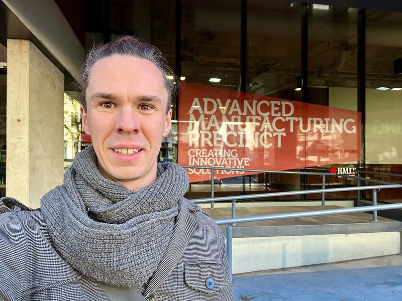 Christopher Taudt steht vor einem Bürogebäude. Im Schaufenster ist ein Transparent mit der Aufschrift "Advanced Manufacturing Precinct" angebracht. 
