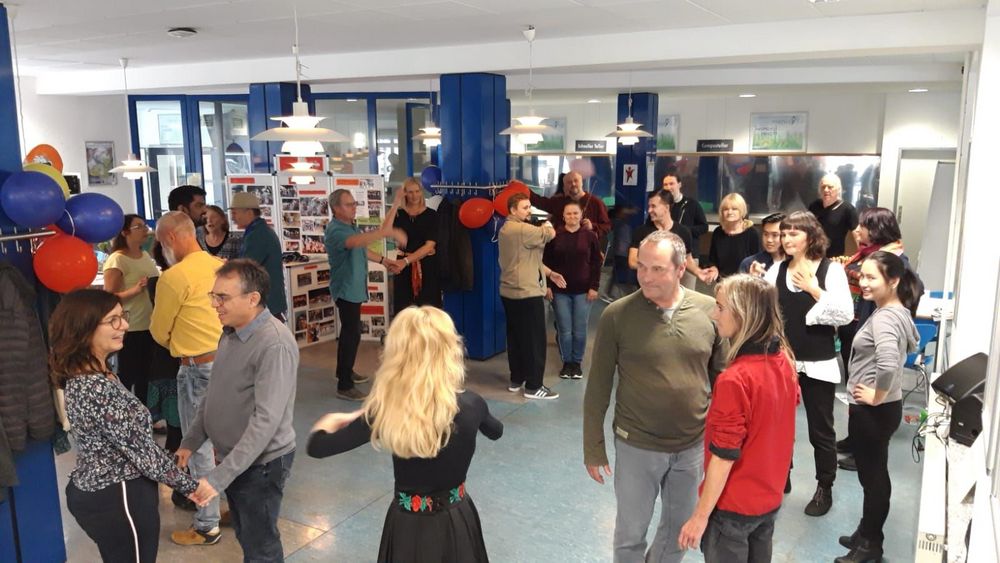 In einem Raum mit belauen Säulen stehen viele Menschen paarweise und warten auf Anweisungen durch eine Tanzlehrerin, die von hinten zu sehen ist. 