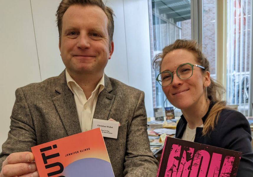 Ein Mann und eine Frau stehen in einem Büro und halten jeweils ein Buch in die Kamera. 