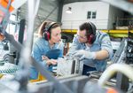 junger Mann und junge Frau in Arbeitsbekleidung und mit Gehörschutz in einer Produktionsumgebung