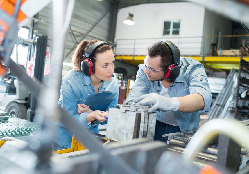 junger Mann und junge Frau in Arbeitsbekleidung und mit Gehörschutz in einer Produktionsumgebung