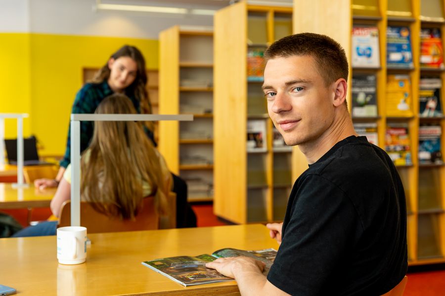 Ein Studierender sitzt in der Hochschulbibliothek und blickt in die Kamera