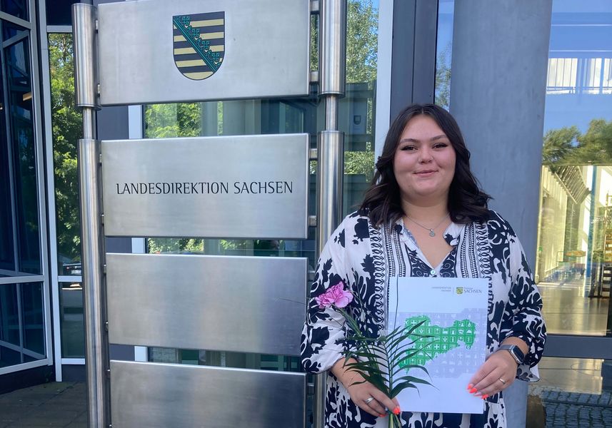 junge Frau mit langen, dunklen Haaren hält glücklich ihr Abschlusszeugnis in die Kamera
