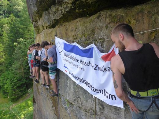 Foto: Mehrere Teilnehmer hängen in einer Felswand mit einem Plakat vom Logo des Hochschulsportzentrum.