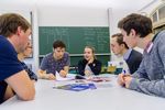 Foto: Eine Gruppe junger Menschen sitzt im Gespräch um einen Tisch, im Hintergrund ist eine beschriebene Tafel.