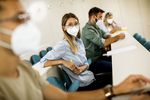 Foto: Eine junge Frau mit Maske sitzt in einer Reihe in einem Hörsaal. Mit einigem Abstand sitzen weitere Studierende.