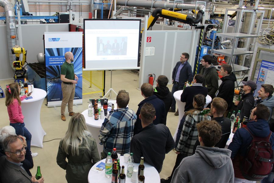 Präsentation in einer Laborhalle. Referenz spricht vor einer Leinwand, Zuhörer stehen und hörenb zu