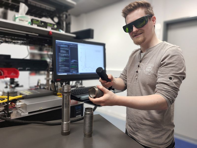 Mann steht im Labor, in der Hand ein Werkstück, Lichschutzbrille auf