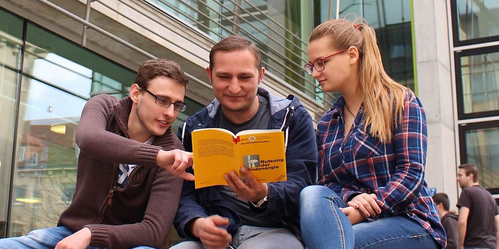 Foto: Drei Studierende sitzen vor der Hauptbibliothek und schauen gemeinsam in ein Buch zur Nutzung der Windenergie.
