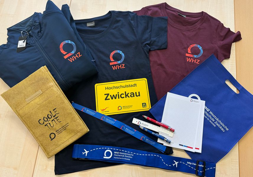 T-Shirts, Jacken, Frühstückbrettchen und weitere Werbeartikel mit dem logo der Westsächsischen Hochschule Zwickau liegen auf einem Tisch. 