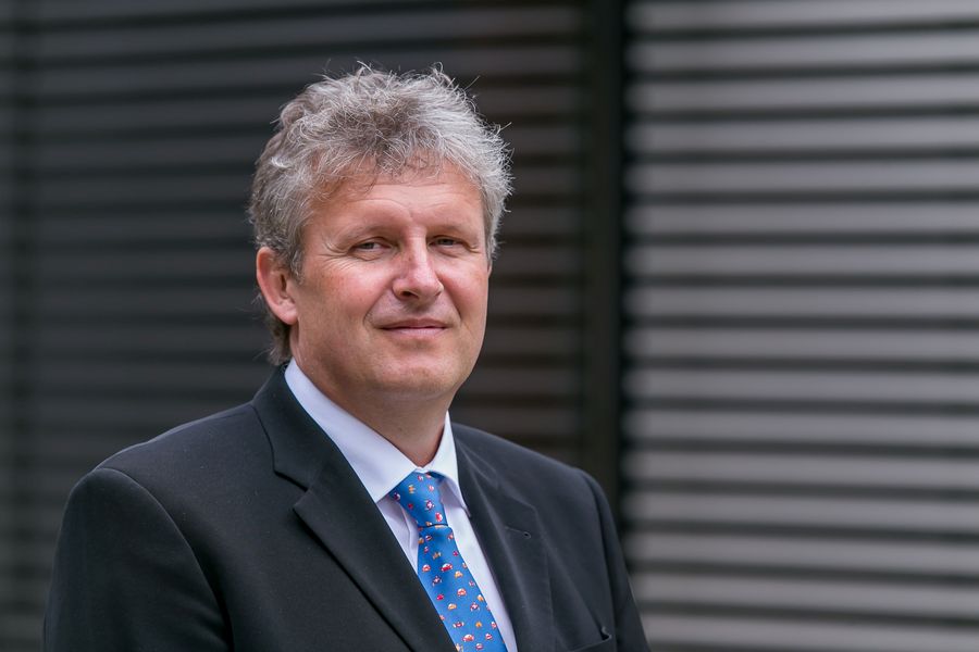 Foto: Portrait eines Mannes, der vor einer grau-schwarzen Wand steht. 