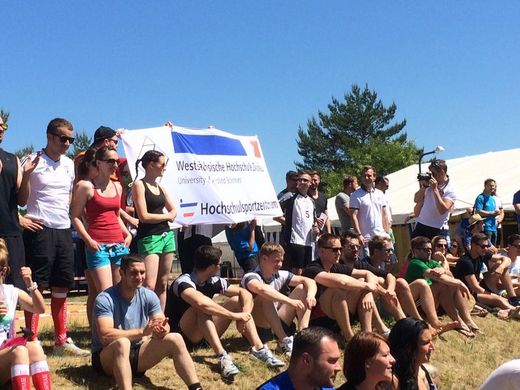 Foto: Teilnehmer des Hochschulsport sitzend und stehend auf einer Wiese mit dem Banner des Hochschulsportzentrum.