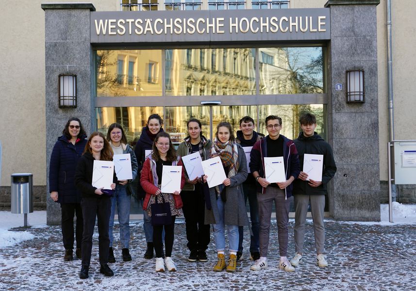 Schülerinnen und Schüler präsentieren ihre Teilnahmeurkunden. Im Hintergrund ein Eingangsportal aus Glas mit dem Schriftzug "Westsächsische Hochschule" 