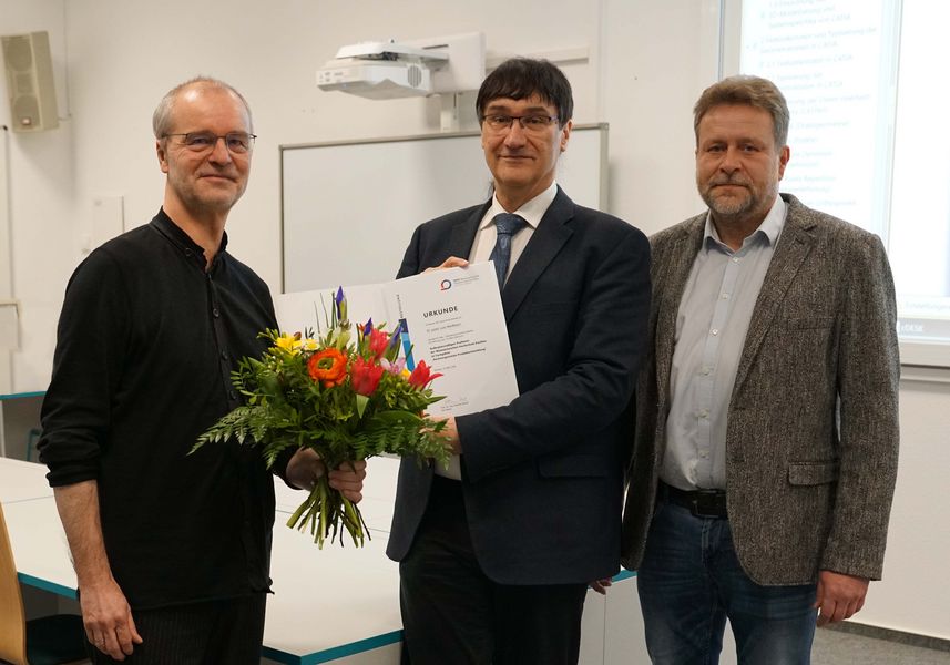 Zwei Männer übergeben die Ernennungsurkunde und den Blumenstrauß für eine außerordentliche Professur an einen dritten Mann