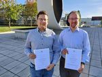 Foto: Zwei Männer stehen vor dem Beton-Looping der WHZ und halten ihre Promotionsurkunden vor sich.
