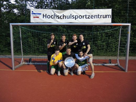 Foto: Gruppenbild. 7 Männer einer Sportgruppe stehen mit Siegerschale in einem Fussballballtor.