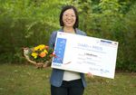 Foto: Yuhong Liu steht im Grünen und blickt in die Kamera. In der Hand hält sie einen Blumenstrauß und einen symbolischen Scheck.