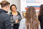 Drei Studieninteressenten hören einer Person beim Sprechen vor einem Plakat zu