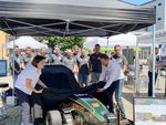 EIne Frau und ein Mann ziehen ein Tuch von einem Rennwagen, im Hintergrund stehen mehrere junge Männer vom Racing Team.